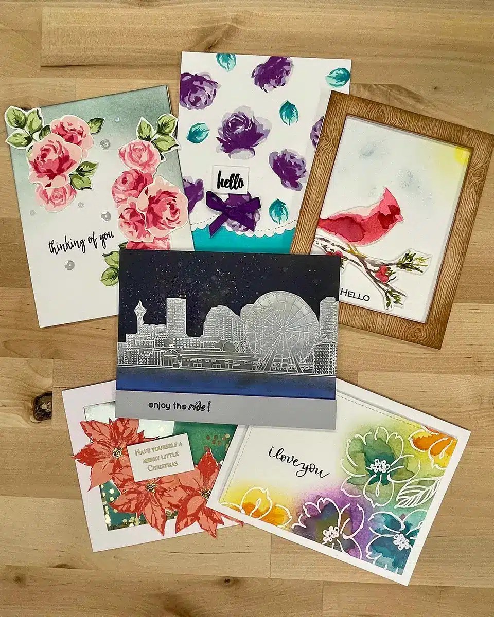 A collection of six handmade greeting cards is laid out on a wooden surface. The cards, showcasing expert ink blending, feature various designs including flowers, a cityscape, and a bird.