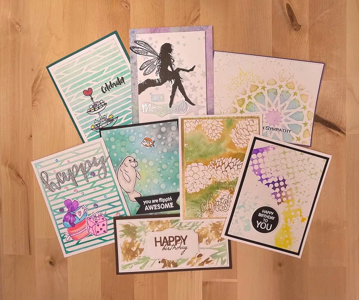 A collection of eight handmade greeting cards arranged on a wooden surface. The cards feature various designs created using stencils, boasting vibrant colors and heartfelt messages like "Happy Birthday," "Celebrate," and "Happy Christmas." These DIY Cards make any occasion special.