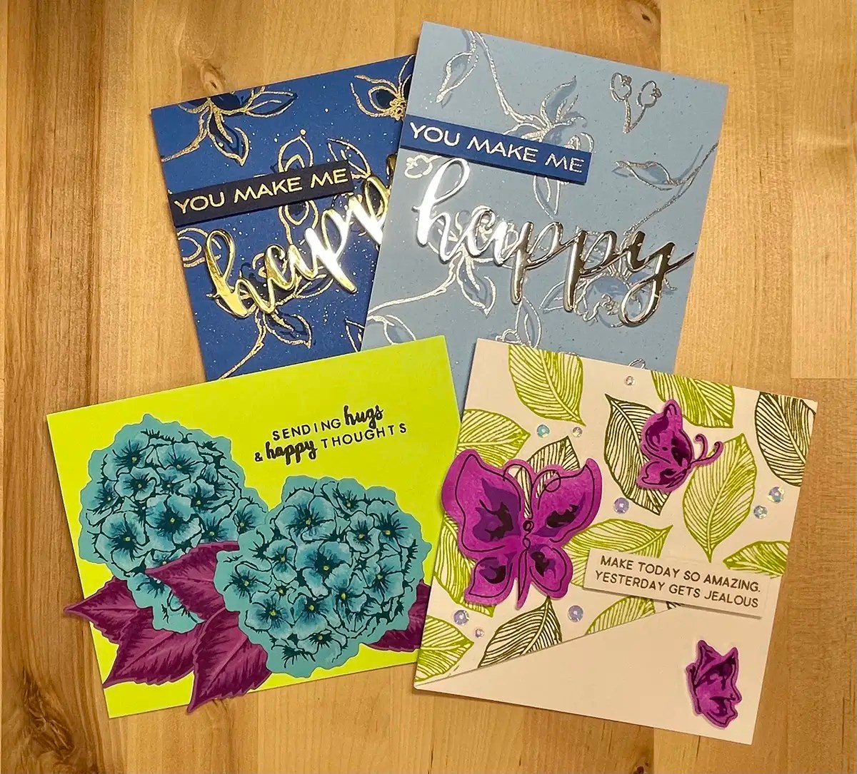 Four cards displayed on a wooden surface, each featuring different designs and messages: "You Make Me Happy," "Sending Hugs & Happy Thoughts," and "Make Today So Amazing Yesterday Gets Jealous." Add a creative twist to your greetings with these vibrant designs.