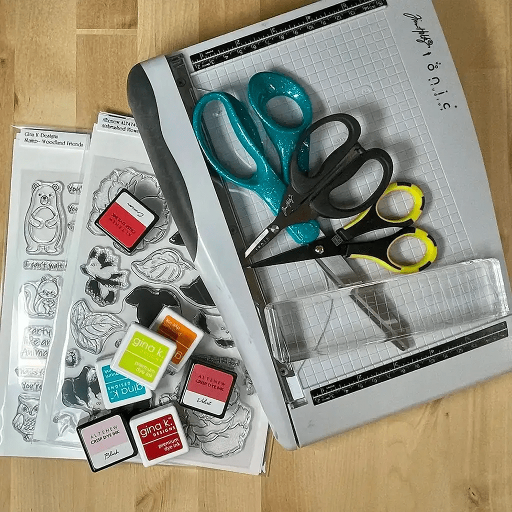 A cardmaking startup tool kit featuring a cutting board, two pairs of scissors, stamp sheets with animal designs, and several small ink pads displayed on a wooden surface.