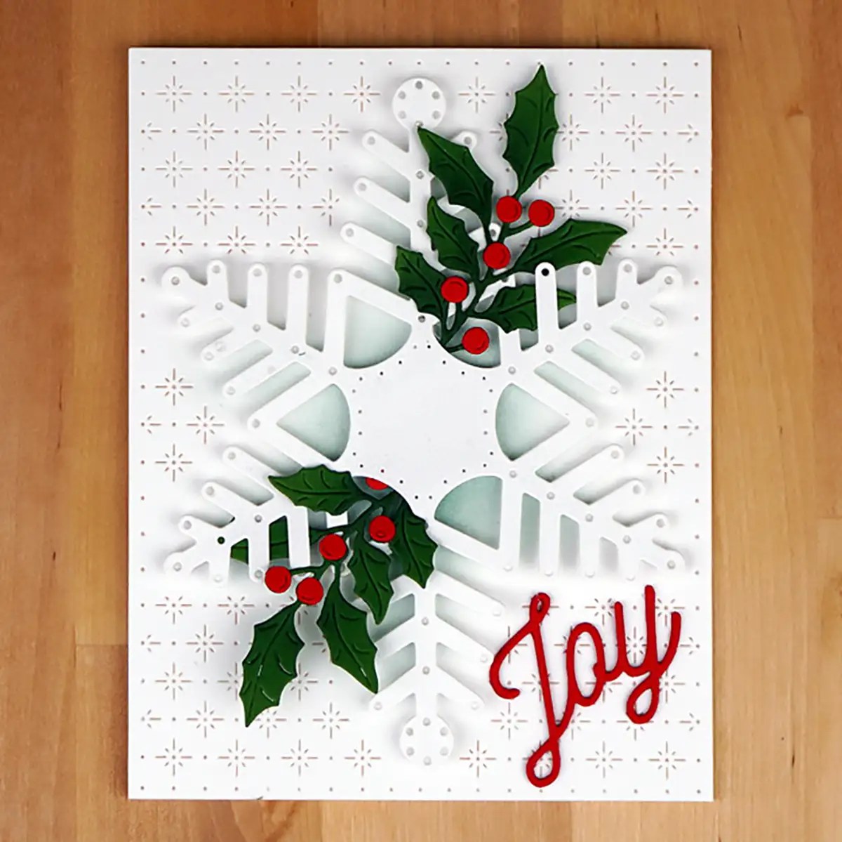 A white card from the Spellbinders July 2023 Monthly Clubs collection features a cut-out snowflake design, adorned with holly and red berries, and the word "Joy" written in red at the bottom right corner.