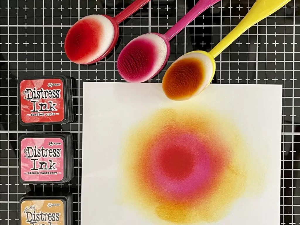 Three blending brushes with colored inks and three distress ink pads sit next to a piece of white paper with a blended circle of red, pink, and yellow ink on a grid mat—perfect tools for creating jaw-dropping cards.