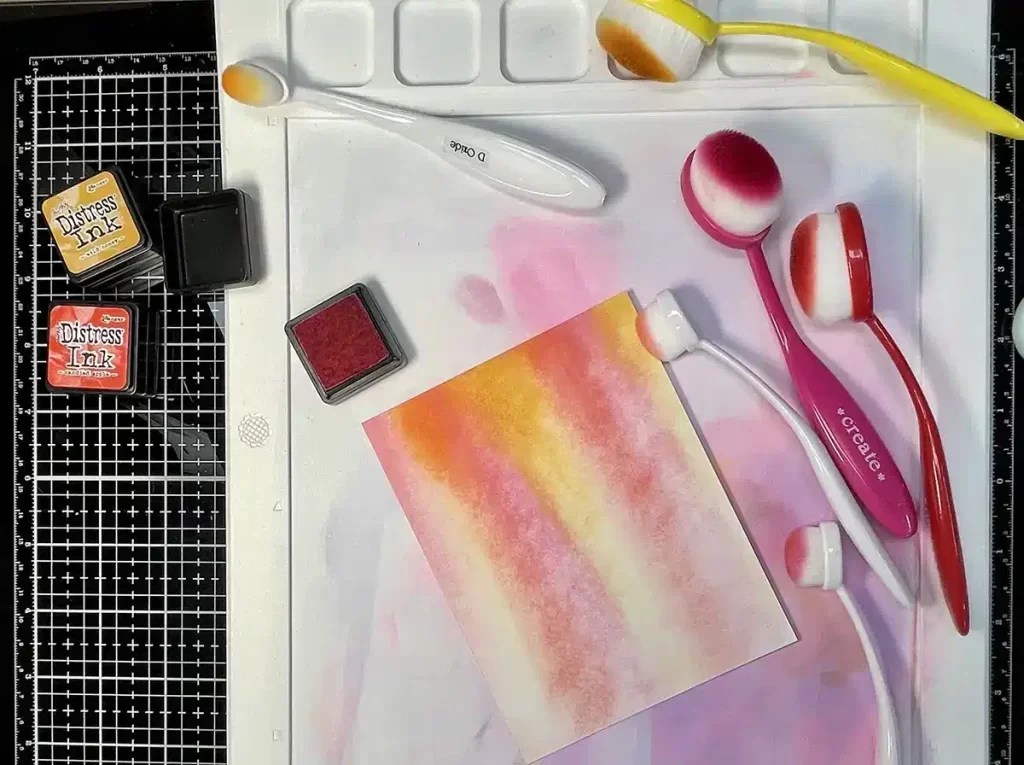 A crafting setup with blending brushes and ink pads on a grid mat, featuring a colorful gradient piece of paper in shades of pink, yellow, and orange—perfect for creating jaw-dropping cards.