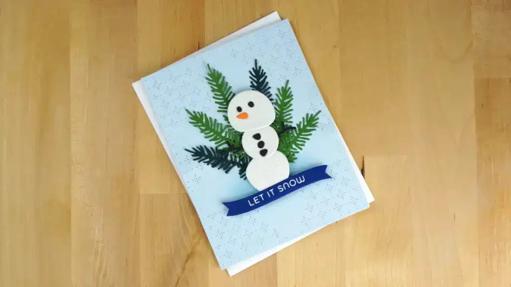 A Winter Greetings card features a cheerful snowman with buttons and green foliage arms, set against a light blue background adorned with snowflake patterns. The Snowman Card includes a blue ribbon with the text "LET IT SNOW.