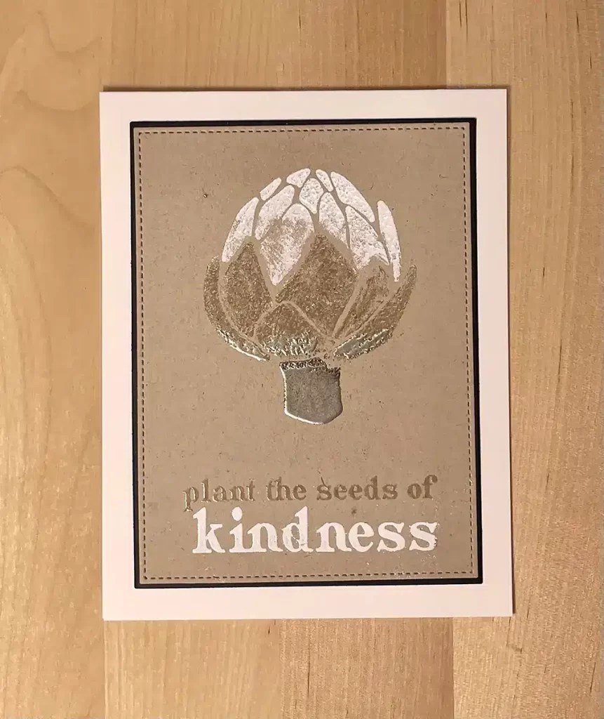 A DIY card with a picture of a flower and the text "plant the seeds of kindness" printed on it, enhanced with heat embossing. The card is placed on a wooden surface.