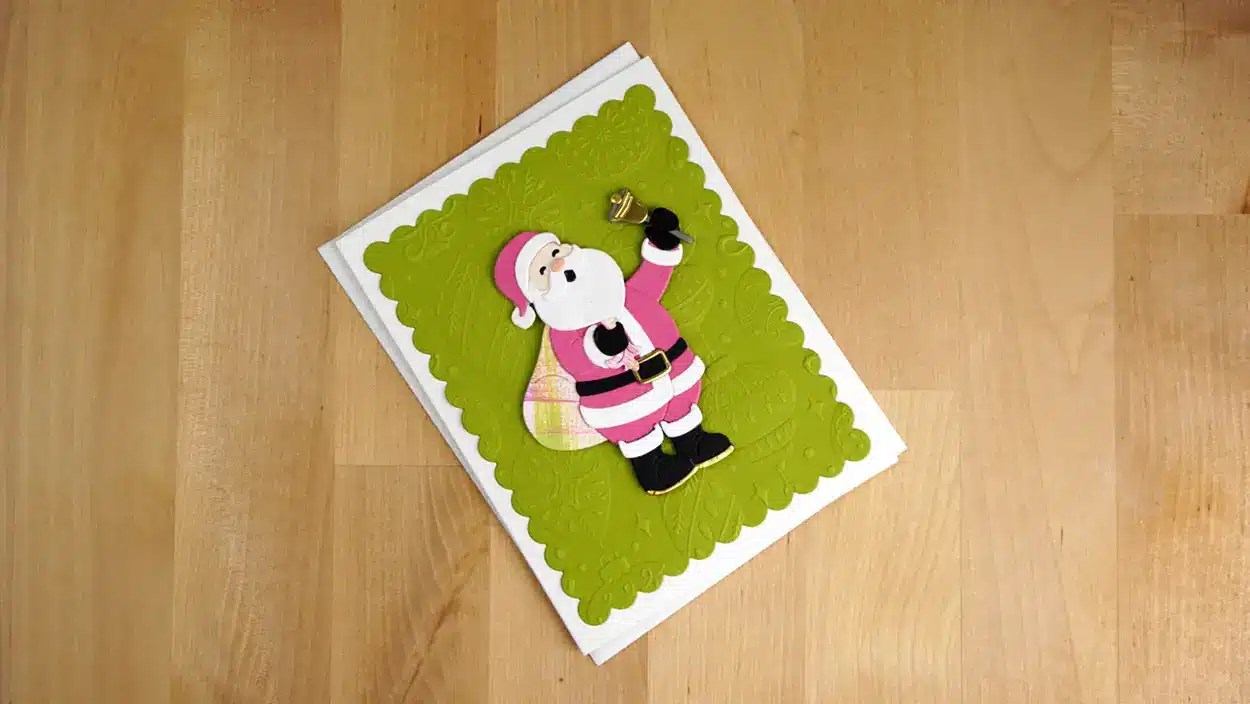 A Christmas greeting card featuring a Santa Claus design on a green scalloped background, with Santa holding a bell and carrying a plaid bag. The festive cards are perfectly placed on a wooden surface, bringing holiday cheer, even in July.