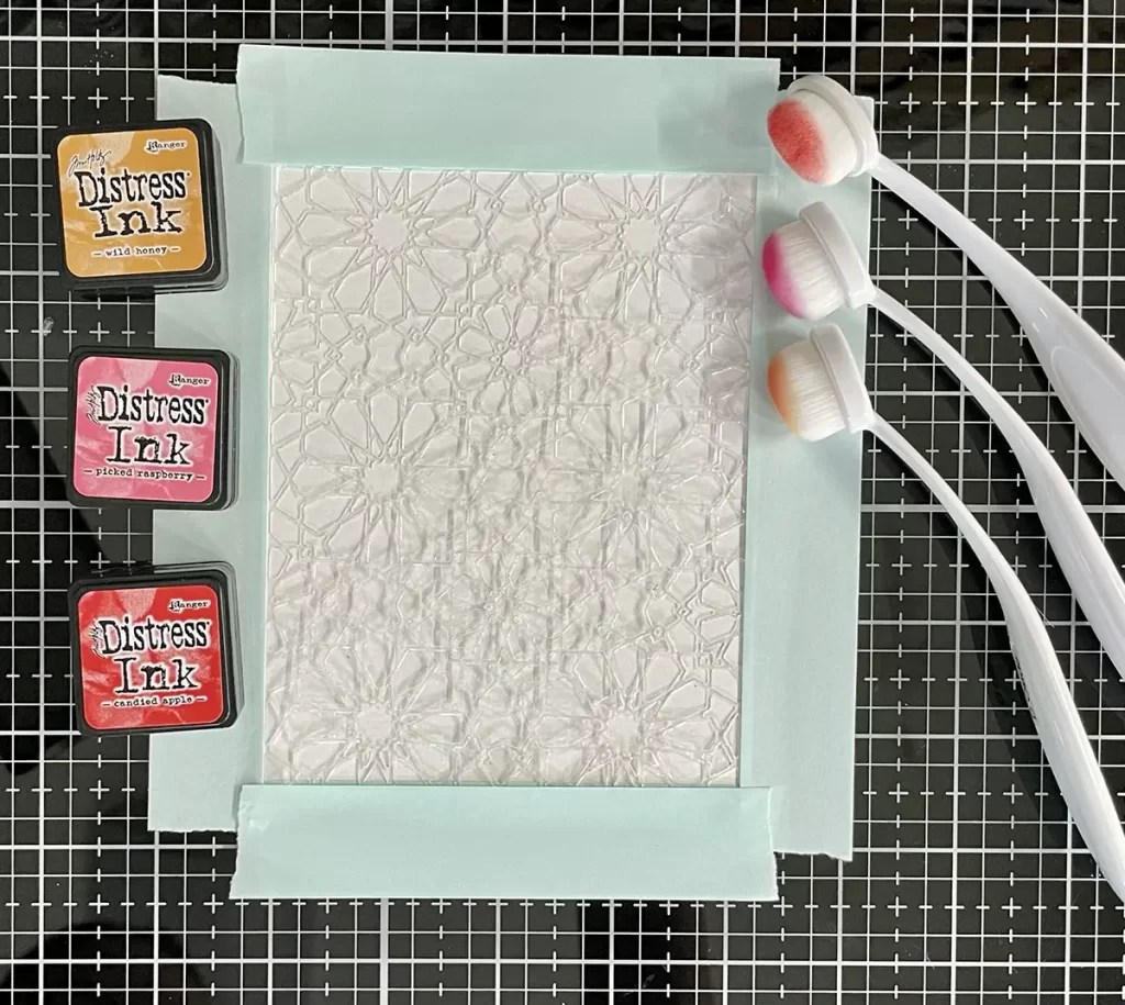 A sheet of paper taped to a grid surface is surrounded by three distress ink pads and three blending brushes. Perfect for DIY card making, the paper's floral pattern promises jaw-dropping cards that stand out.