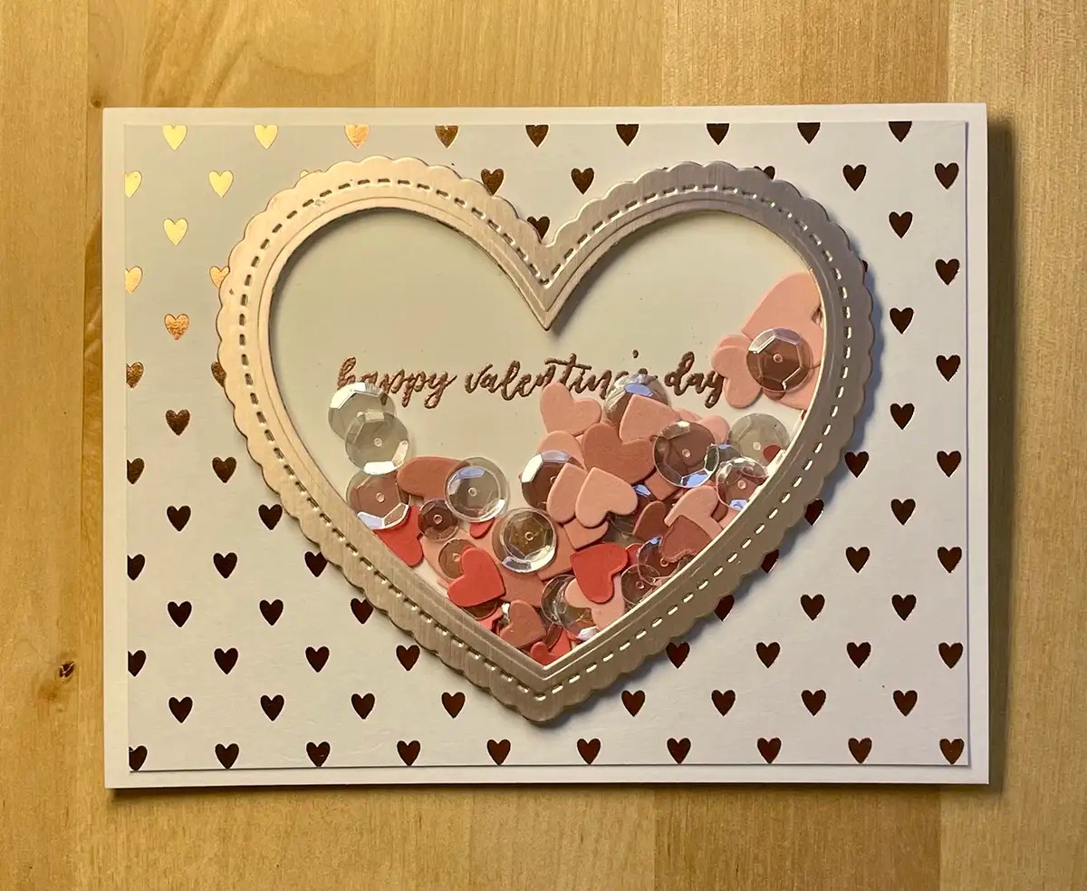 Greeting card with a heart-shaped window filled with small heart and sequin confetti, surrounded by a pattern of tiny hearts, and the text "happy valentine's day" inside. Perfect for those who love creating DIY Valentine Cards.