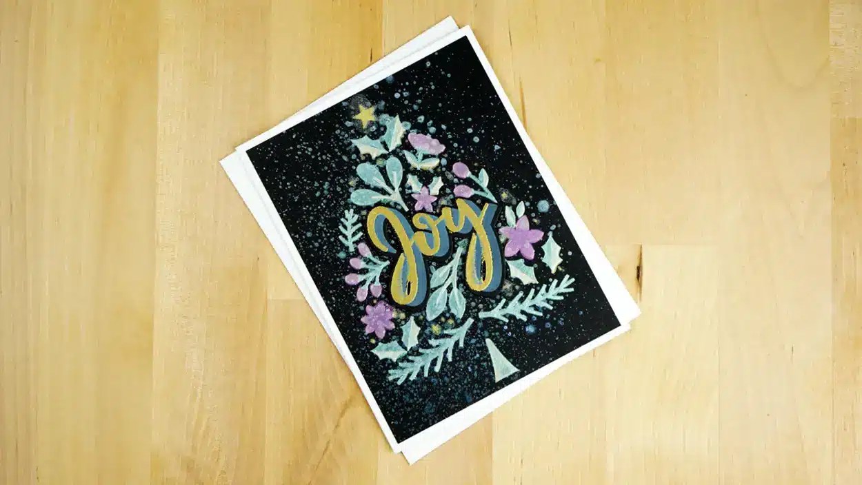 A greeting card with the word "Joy" written on the front, featuring a tree design with colorful decorations, lies on a wooden surface—perfect for celebrating Christmas in July. Available in a set of 8 cards.