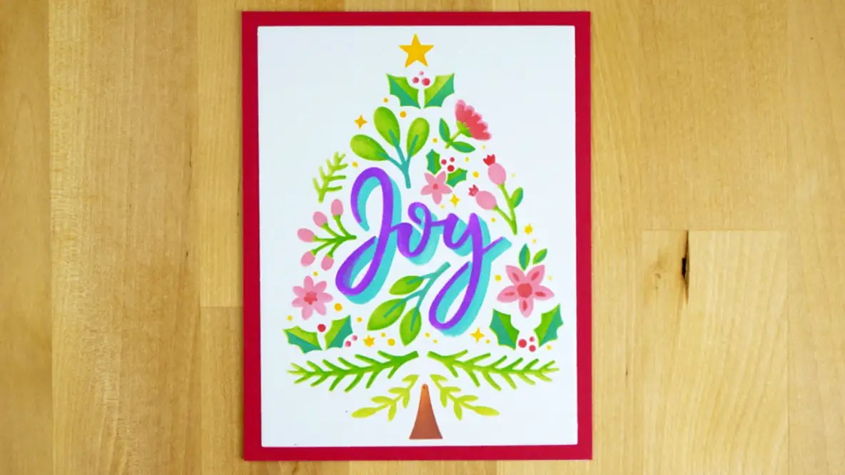 A festive card featuring a Christmas tree design with colorful flowers and leaves, and the word "Joy" written in purple in the center. Perfect for celebrating Christmas in July, this delightful card is topped with a yellow star.