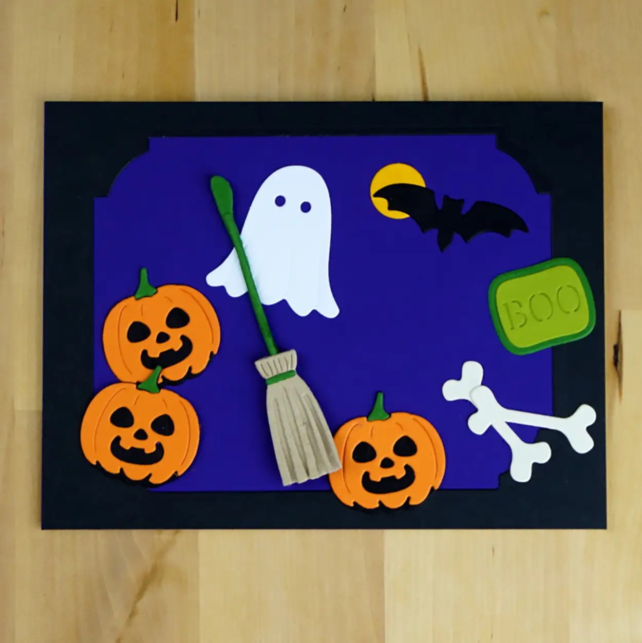 A Halloween-themed craft from the Spellbinders Sept23 Monthly Club Kits, featuring a ghost, three jack-o'-lanterns, a broomstick, a bat, bones, and a "BOO" sign on a purple and black background.