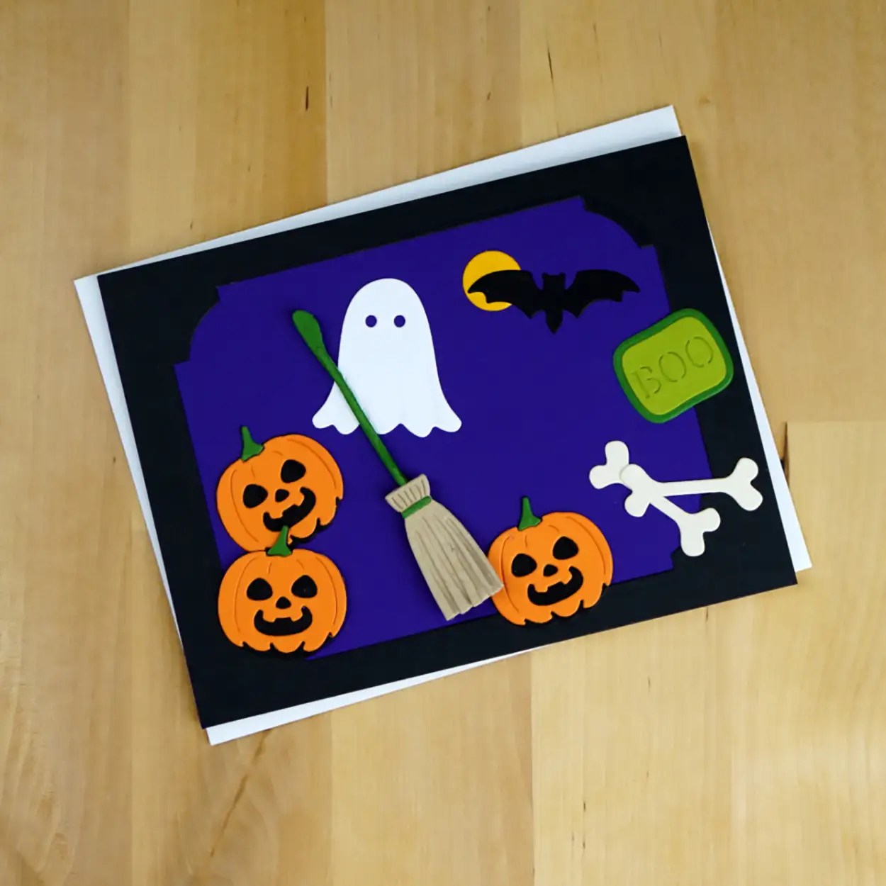 A September 2023 Spellbinders Monthly Club Kits Halloween-themed card is decorated with pumpkins, a ghost, a broom, a bat, a green 'BOO' sign, and two bones against a purple background.