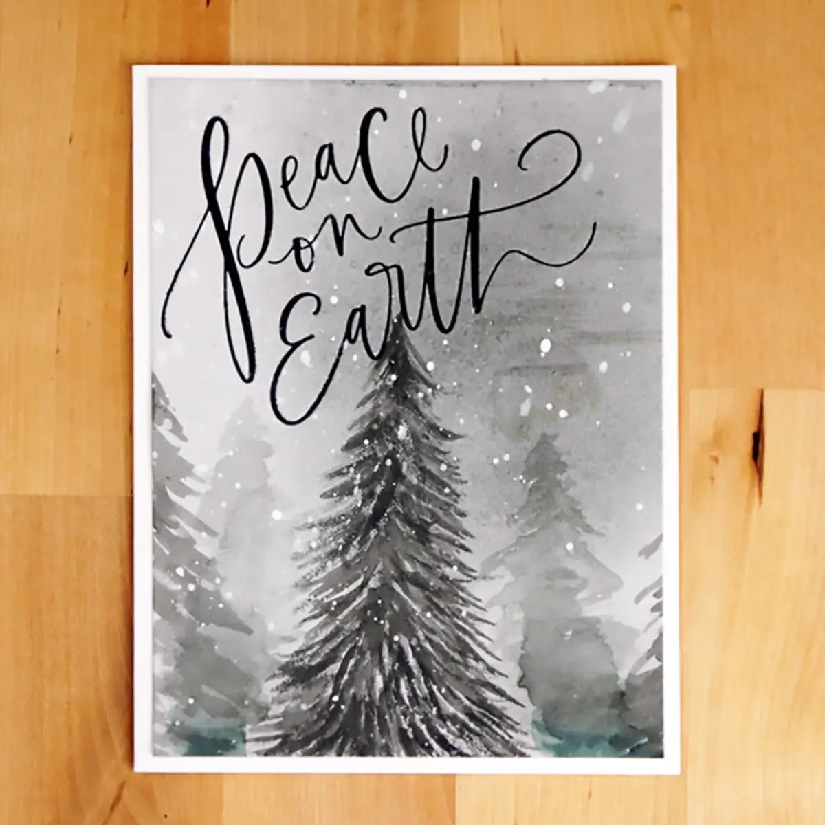 A framed picture lies on a wooden surface, featuring a snowy forest scene with the phrase "Peace on Earth" written in cursive at the top. Perfect for BetterPress Cards to help spread cheer this holiday season.