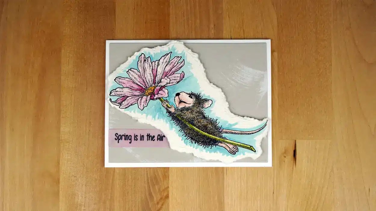 A drawing of a House Mouse holding a pink flower graces the card with the text "Spring is in the air." Part of the Spring Collection, this charming card rests on a wooden surface.