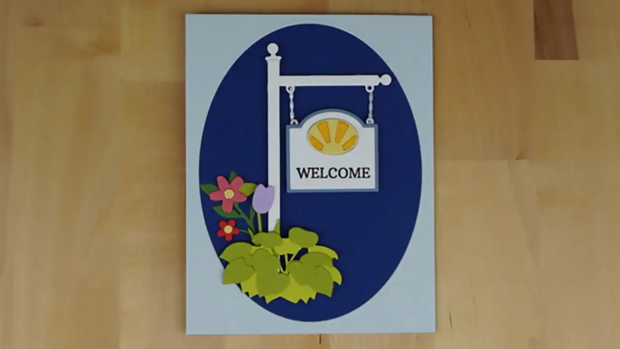 A decorative sign with the word "Welcome" inside an oval frame, accompanied by colorful paper flowers, displayed against a wooden background reminiscent of country roads.