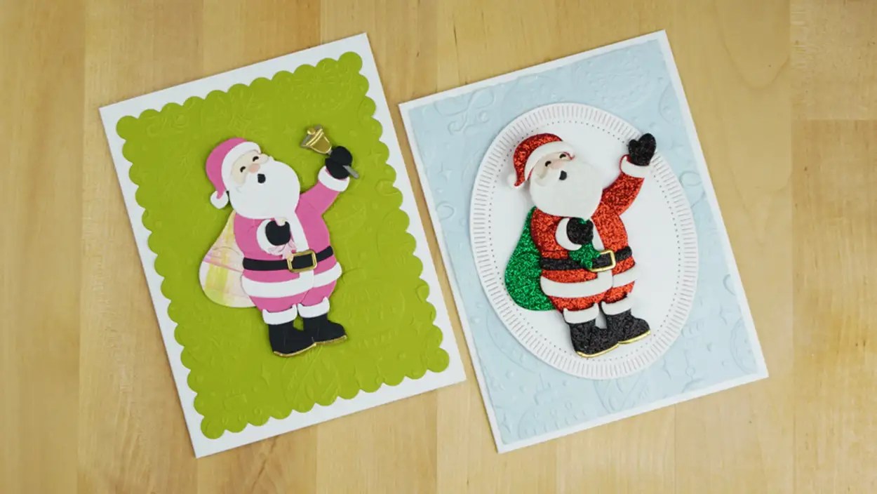 Two Christmas cards featuring Santa Claus—one in a pink suit with a bell on a green background, and the other in a red suit waving on a blue background—lie on a wooden surface. These festive cards bring a touch of Christmas in July to your celebrations.