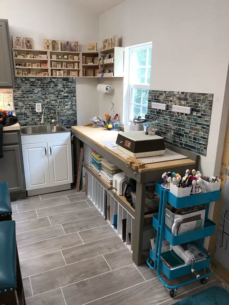 A small, organized crafting workspace with a table, Stamp-N-Storage bins, and a wall-mounted shelving unit. A window provides natural light, and a sink is nearby. A blue cart holds various crafting supplies for the best storage experience.