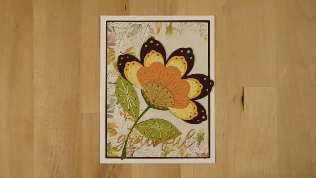 A greeting card with a large flower illustration in autumn-colored hues and the word “grateful” written at the bottom, placed on a wooden surface—perfect for DIY crafts enthusiasts.