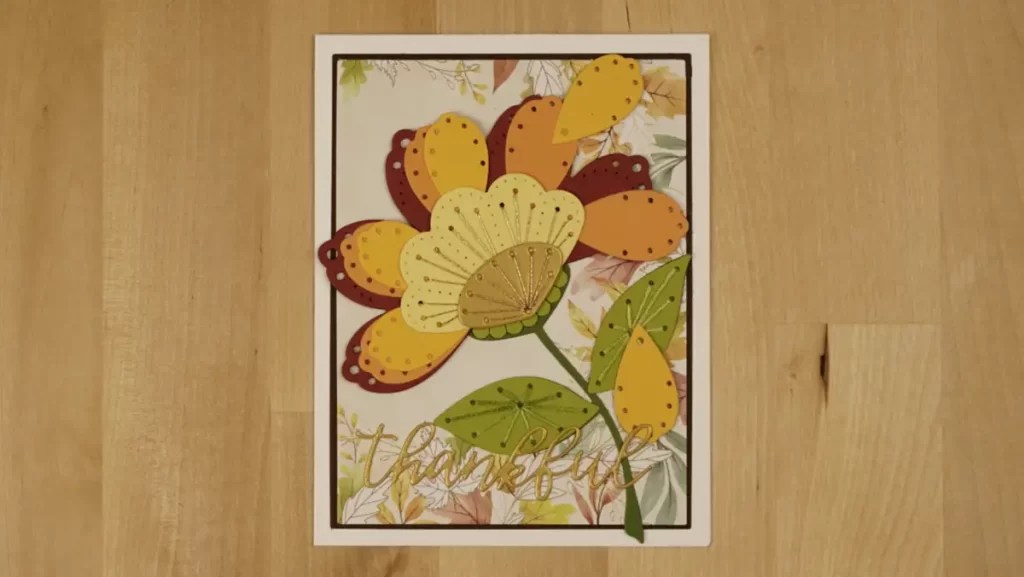 A handcrafted card featuring a large flower with yellow, orange, and red petals, green leaves, and the word "thankful" at the bottom in gold script. The background has a floral pattern, making it perfect for autumn crafts enthusiasts who love stitching cards.