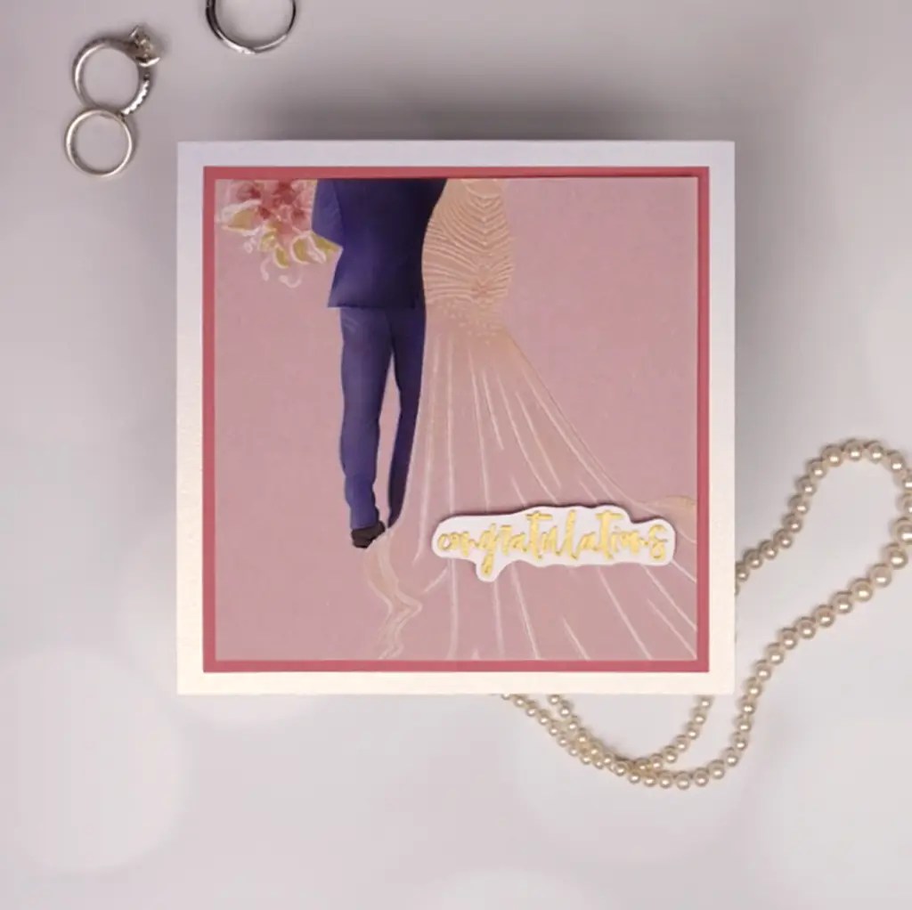 A square, pink wedding card with an illustration of a couple. The woman is wearing a wedding dress, and the man is in a suit. The word "congratulations" is written below. Rings and a pearl necklace are nearby. Ideal for giveaways, this wedding card adds a touch of elegance to any special day.