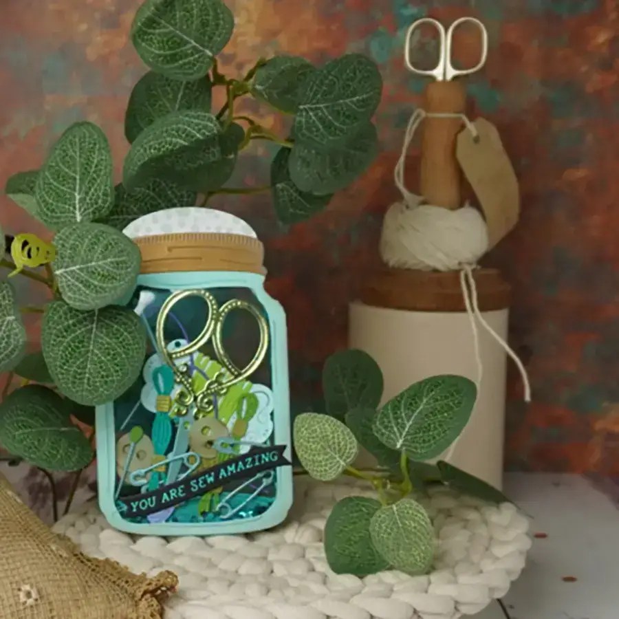 A decorative jar with sewing accessories and a tag that reads "You are sew amazing," surrounded by green plants. In the background, a pair of scissors and a spool of white thread add charm. Perfect for any crafting corner, this Apr24C edition is both functional and inspiring.