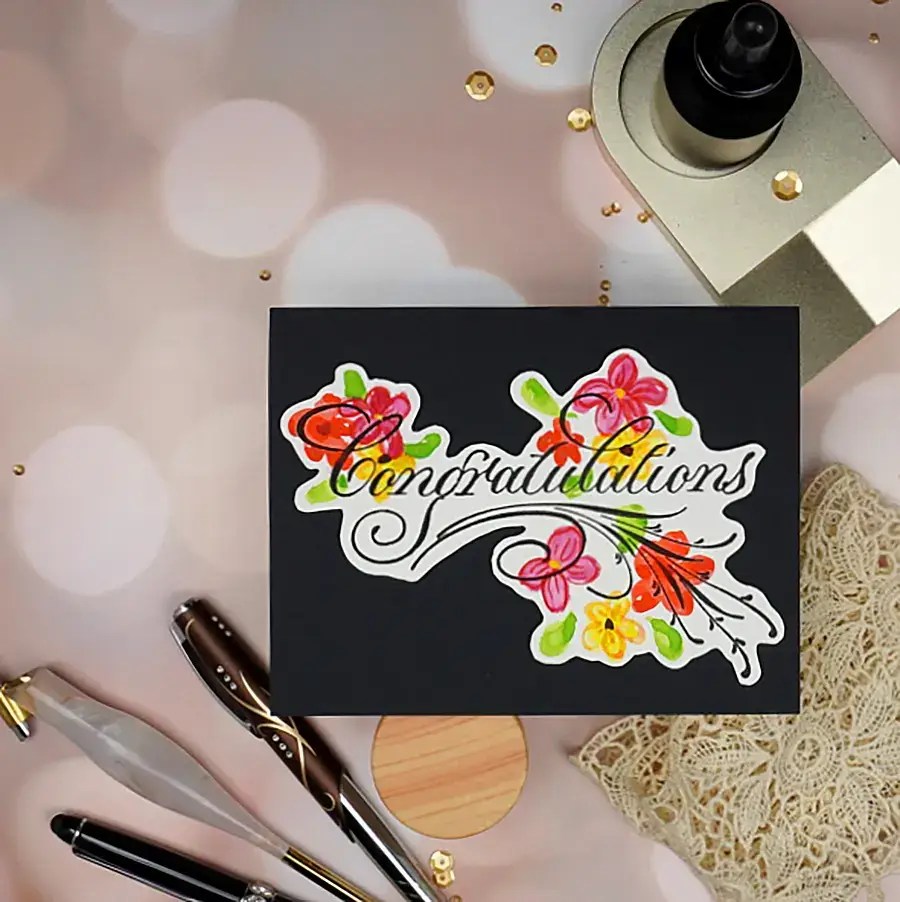 A "congratulations" card with colorful flowers on a black background, surrounded by a fountain pen, dip pen, ink bottle, lace cloth, and scattered gold sequins. The elegant Copperplate script adds a touch of sophistication to the celebratory tone.
