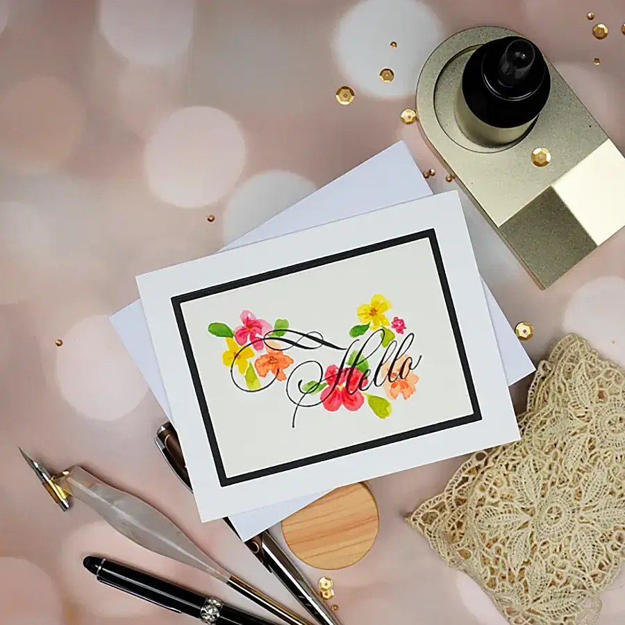 A card with colorful flowers and "Hello B" elegantly written in Copperplate script, placed on a desk with a pen, lace doily, ink bottle, and white envelopes, all adorned with golden confetti scattered across the surface.