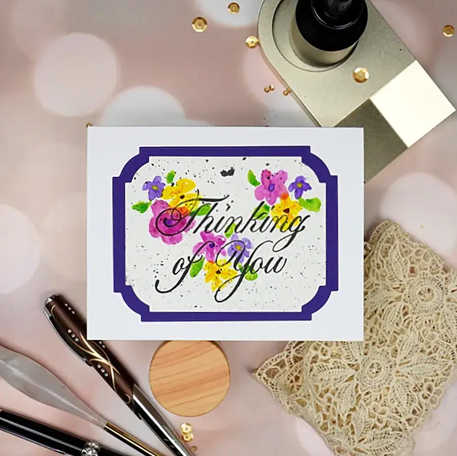 A handmade greeting card with "Thinking of You" text in elegant Copperplate script, surrounded by floral decorations. The card is placed on a table with various greeting stationery items and a lace napkin nearby.