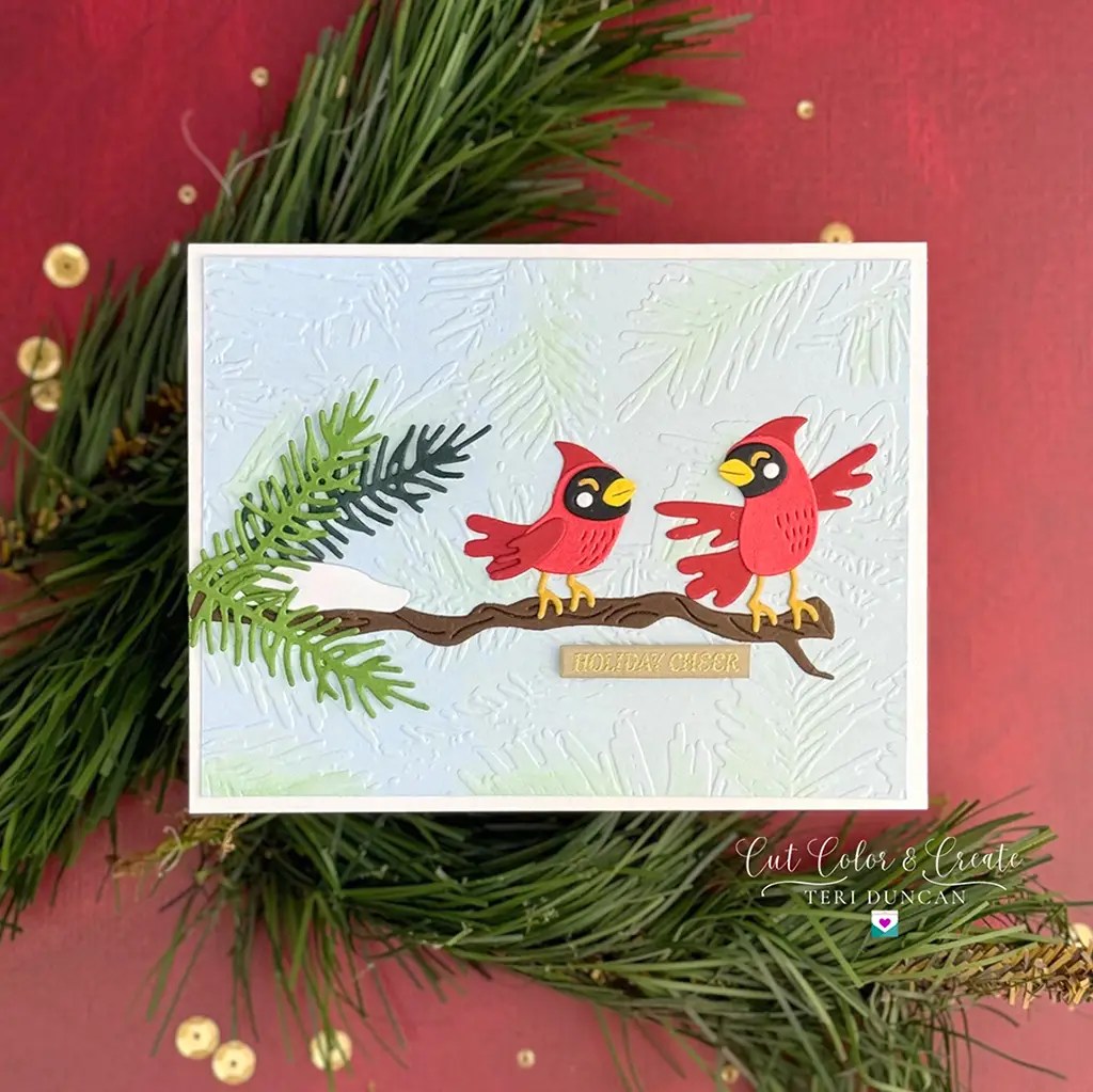 A Spellbinders greeting card with two red birds perched on a branch, set against a textured background and framed by pine branches. The bottom of the card reads "HOLIDAY CHEER". Don’t miss out—get yours through our Monthly Clubs starting July 2024!