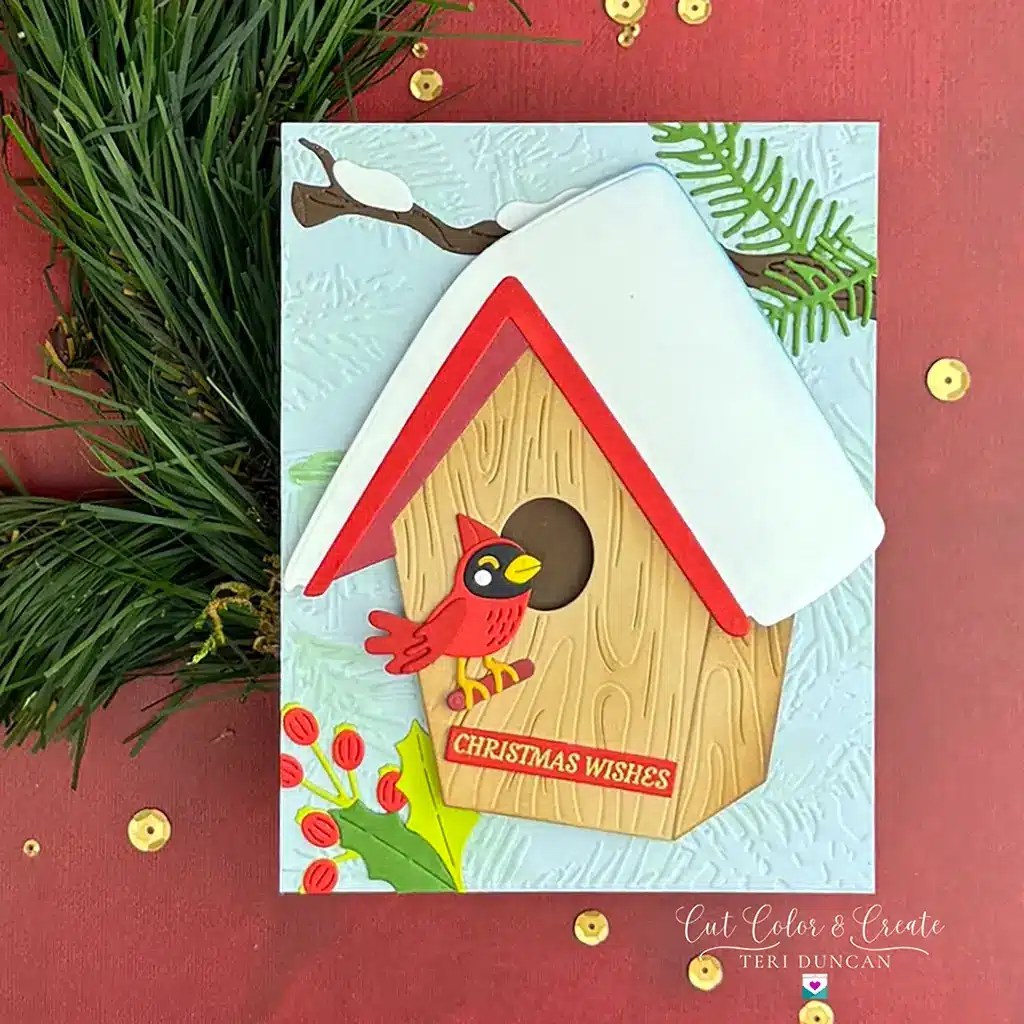A handmade card from Spellbinders' July 2024 Monthly Clubs features a red-roofed birdhouse with a red bird, pine leaves, and "Christmas Wishes" text. Pine branches are in the background.