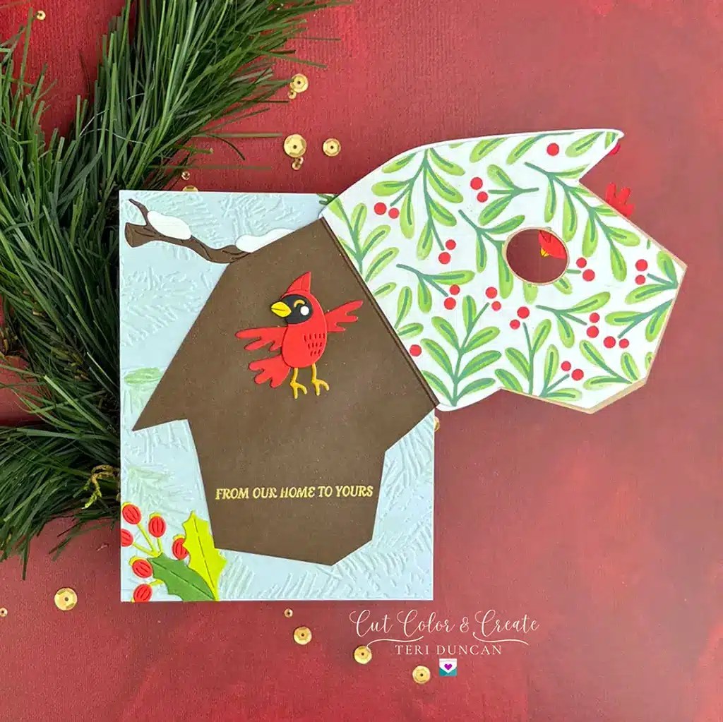 A handmade holiday card by Spellbinders, decorated with a red cardinal on a birdhouse and green branches with red berries. Text reads, "From our home to yours." The card is placed on a red surface with pine sprigs. Look out for it in our monthly clubs, available from July 2024.