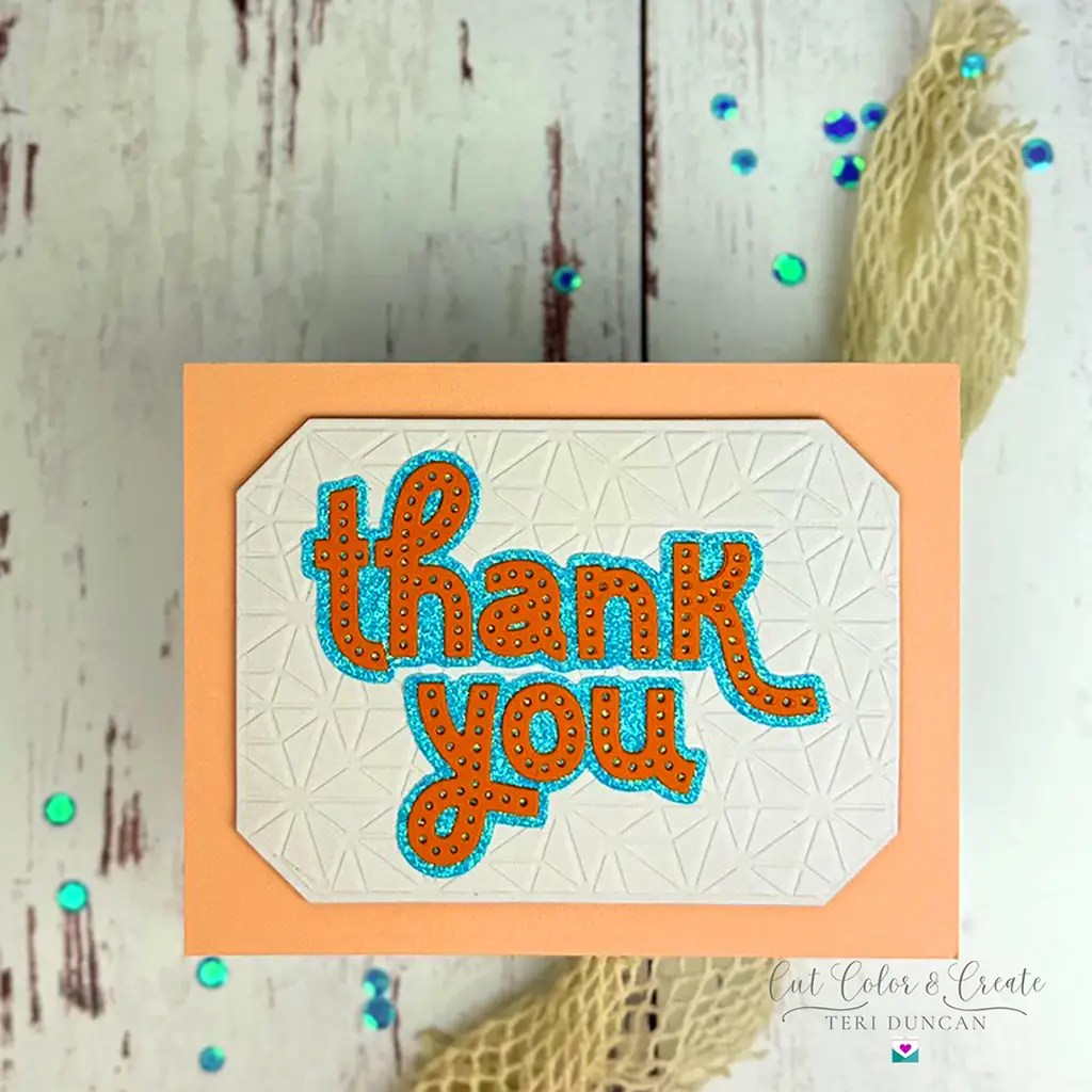 A handmade card with the text "thank you" in orange and blue letters on a textured white background, set against an orange border. The card is placed on a white wooden surface with scattered blue sequins.