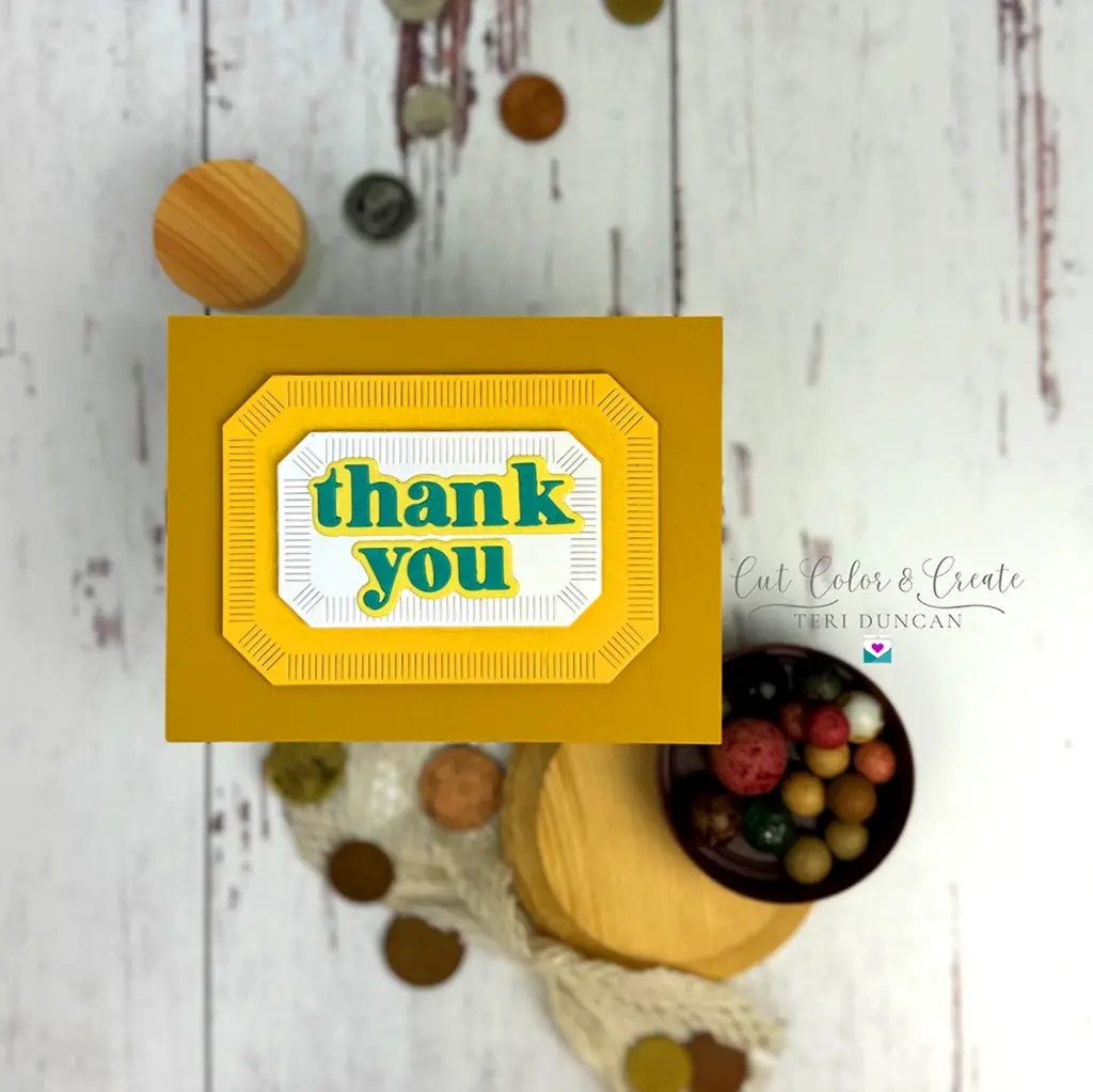 A yellow thank you card with embossed edges sits on a white wooden table. Several small round objects and textured fabric are placed around it.