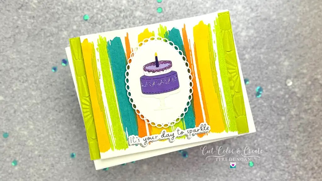 A handmade card with a purple cake on a stand, surrounded by colorful vertical brush strokes. Text reads, "It's your day to sparkle made using November 24 Club Kits.