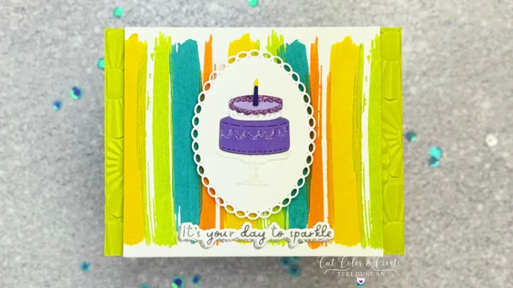 A colorful, handmade card with a purple cake illustration in the center. The background features vertical stripes in green, yellow, and orange. Text reads, "It's your day to sparkle made using November 24 Club Ki.