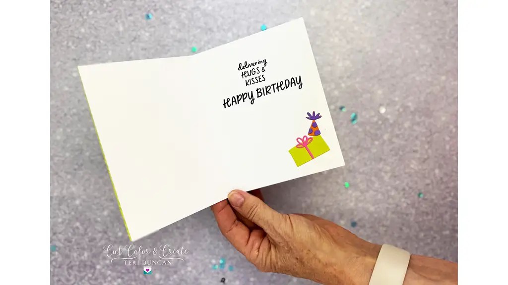 A hand holding an open birthday card on a glittery surface. The card reads, "Sending hugs & kisses. Happy Birthday," with an illustration of a gift and flowers.