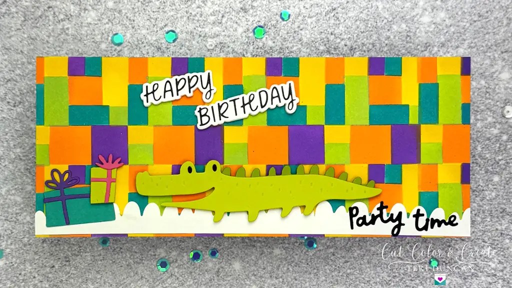 Colorful birthday card with a green crocodile, presents, and text saying "Happy Birthday" and "Party Time." Background features a vibrant woven pattern of orange, green, and purple made using November 24 Club Ki.