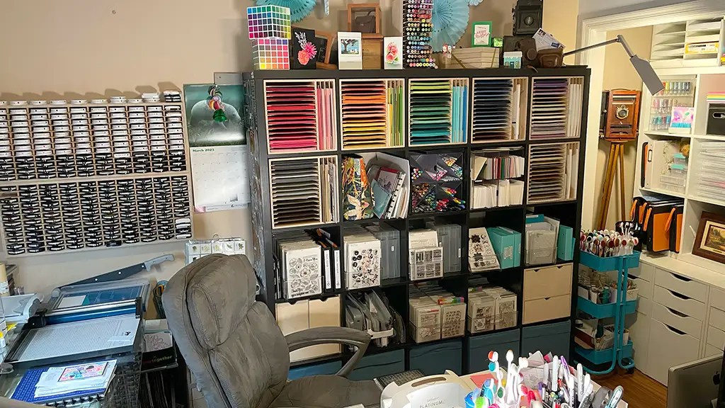 A craft room showcasing impeccable organization hacks, with shelves brimming with colorful paper and supplies. A desk adorned with markers and tools, complemented by a cushioned chair, completes the scene where every organized material inspires creativity.