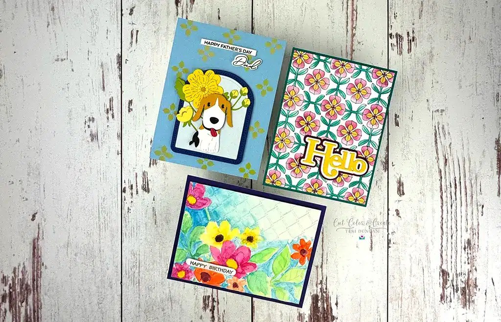 Three handmade greeting cards on a wooden surface feature a Father's Day card with a dog, a floral card with "Hello," and a watercolor floral birthday card, all crafted using Spellbinders from the March '25 Kits.