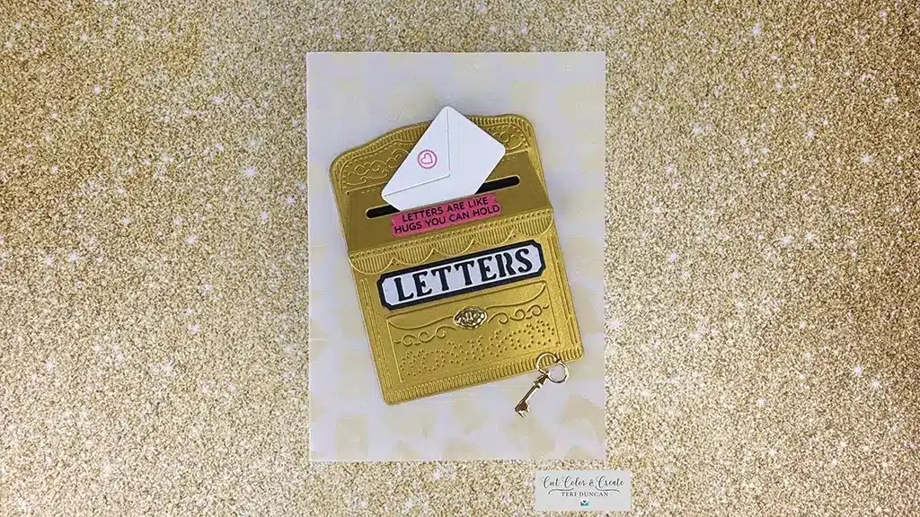 Gold glittery background with a card showing a gold mailbox reading "LETTERS." An envelope inside reads, "LETTERS ARE LIKE HUGS YOU CAN HOLD." A small key embellishment is attached, capturing the charm of an April 25 Club Kits exclusive.