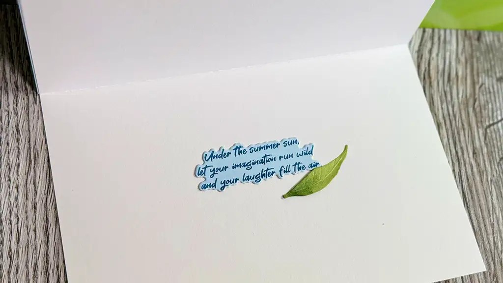 An open Summer Solstice Card on a wooden surface with a small green leaf and handwritten text: "Under the summer sun, let your imagination run wild and your laughter fill the air.