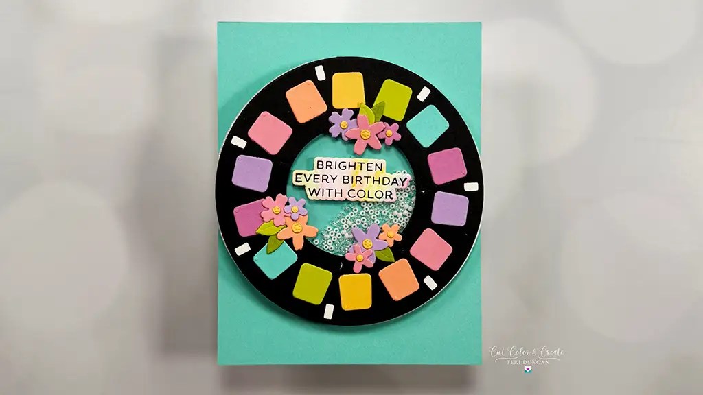 A handmade birthday card featuring a black reel with colorful squares, small flowers, and the message "Brighten every birthday with color" on a teal background—perfect for anyone searching for unique cardmaking ideas. This card is created with Color Reel from Spellbinders Take A Note collection.