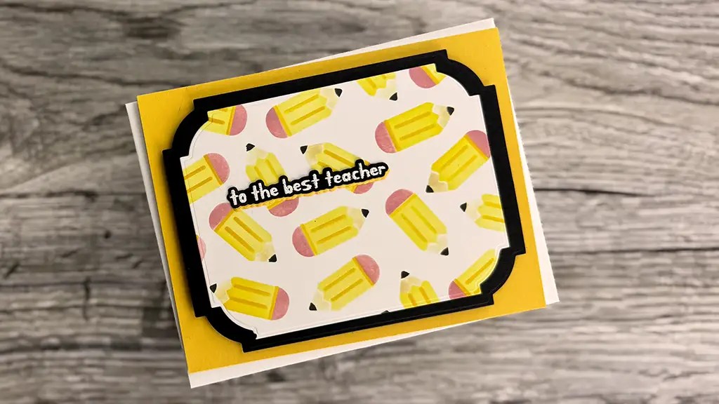 A greeting card with a pattern of yellow pencils and the text "to the best teacher" on the front, perfect for May 2025 or as inspiration for card making and club kits, placed on a wooden surface.
