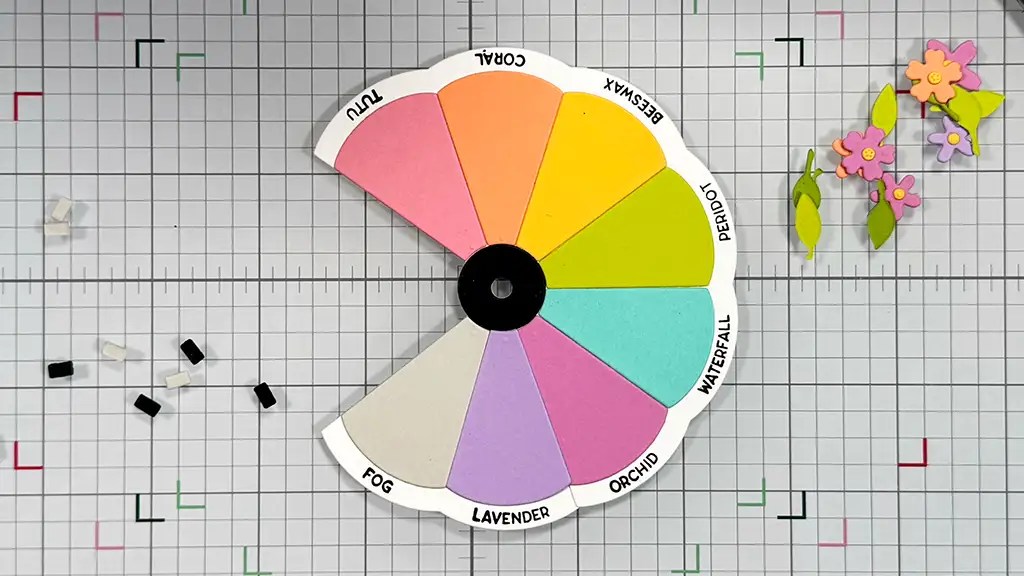Take A Note color palette and cardstock swatches with six labeled segments—Tutu, Coral, Beeswax, Peridot, Waterfall, Orchid, Lavender, and Fog—lies on a grid surface next to paper flowers and small craft pieces, inspiring fresh cardmaking ideas.
