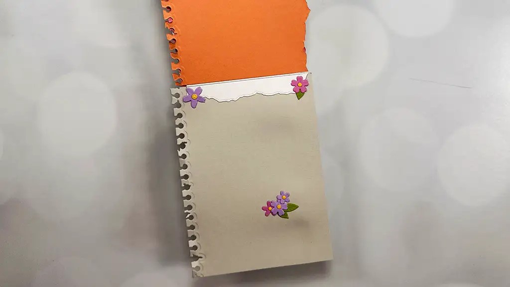 A torn-out orange notebook page, perfect for note cardmaking, is partially inserted into a cream-colored notebook page decorated with purple flower stickers.