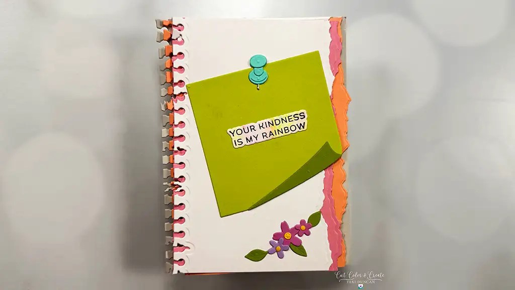 A spiral-bound notebook with a decorated cover featuring a green note that reads “Your kindness is my rainbow,” a blue pushpin illustration, and flower embellishments.This card is made with products from Spellbinders take a note collection.