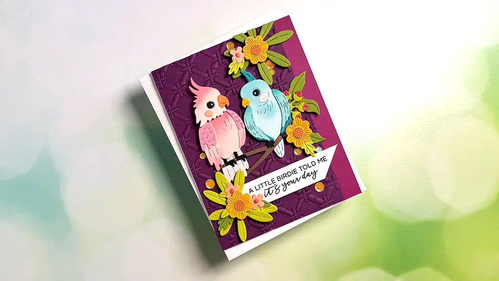 A handmade card from the Cheerful Chirps collection features paper cutouts of a pink and blue bird, yellow flowers, and the message "A little birdie told me it's your day" on a purple background.