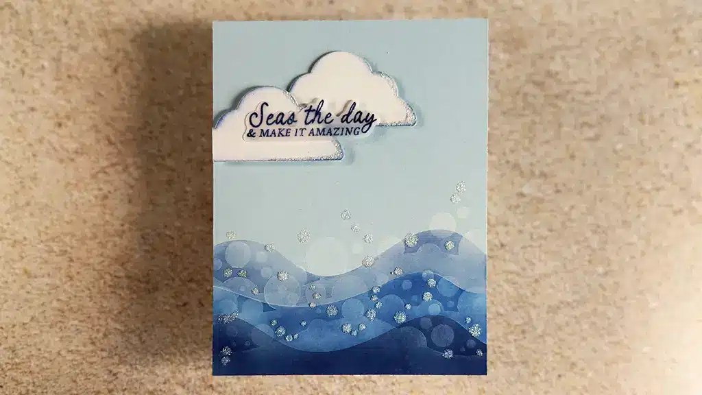 A Nautical Card featuring blue waves, a cloud, and the text “Seas the day & make it amazing” on a sandy background—perfect for Club Kits or coastal-themed occasions. Create this card using products from Spellbinders June '25 club kits.