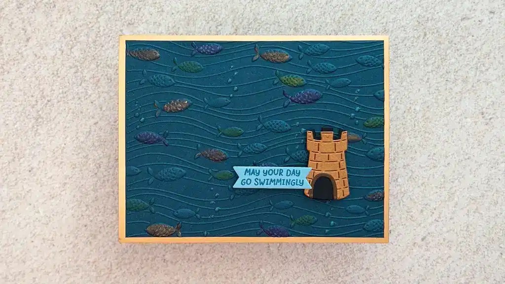 A greeting card with a fish and wave pattern, a small castle illustration, and a message that reads, "May your day go swimmingly. This card is made with products from Spellbinders August '25 kits.