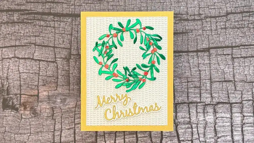 A handmade card with rich texture, showcasing a green foliage wreath and red berries, featuring "Merry Christmas" in gold letters on a textured white background. Created using products from Spellbinders Textures & Folliage collection